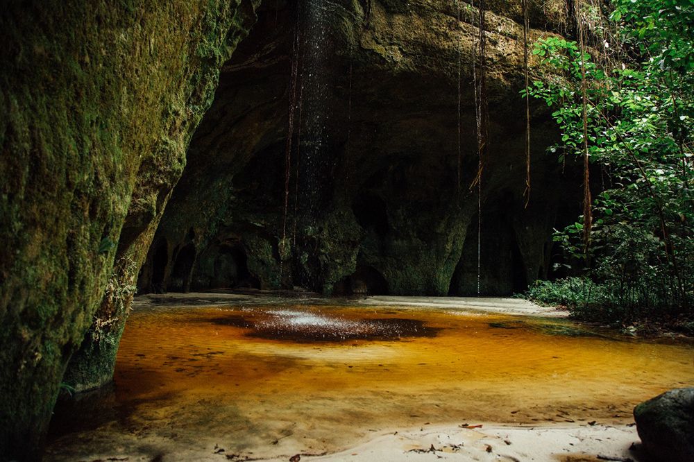 Amber-tinted pool inside a sandstone cave with a waterfall curtain and hanging vines — Gruta da Judéia, Presidente Figueiredo